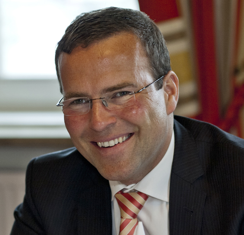 Lächelnder Mann in Anzug und gestreifter Krawatte, mit Brille, sitzt in einem Haus mit roten Vorhängen im Hintergrund.